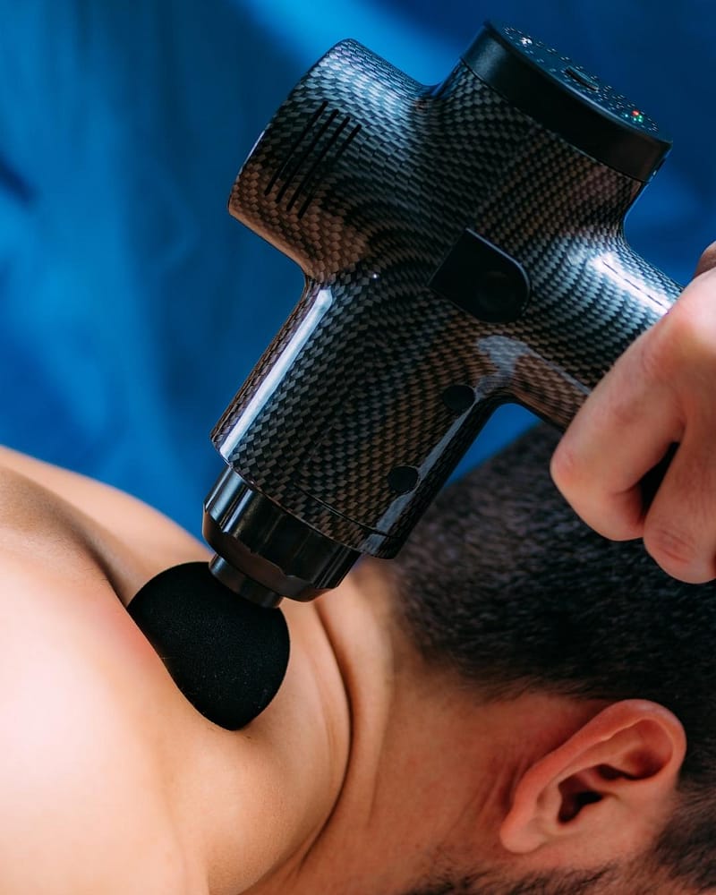 Massage therapist treating desk job neck and shoulder pain with a massage gun on a client’s upper back
